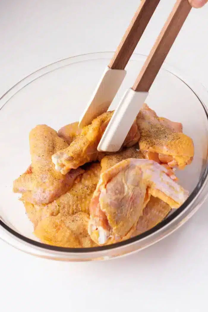 A glass bowl contains several raw chicken wings seasoned with spices.