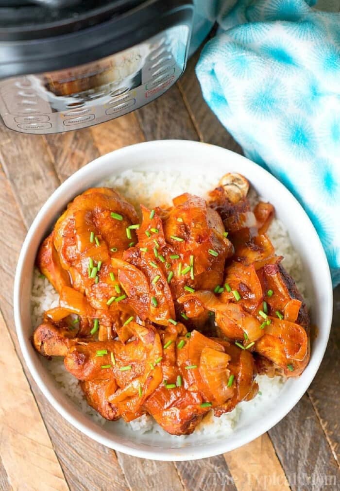 Instant Pot chicken legs are amazing! If you love easy pressure cooker dinners but looking for something your kids will love too chicken drumsticks are simple to make in just 10 minutes. This fall off the bone tender Instant Pot drumstick recipe will be your go to meal for sure! #instantpotrecipes #pressurecooker #chickenlegs #chicken #legs #drumsticks #instantpot #thetypicalmom A bowl of rice topped with saucy instant pot chicken legs and chopped chives sits next to an electric pressure cooker.