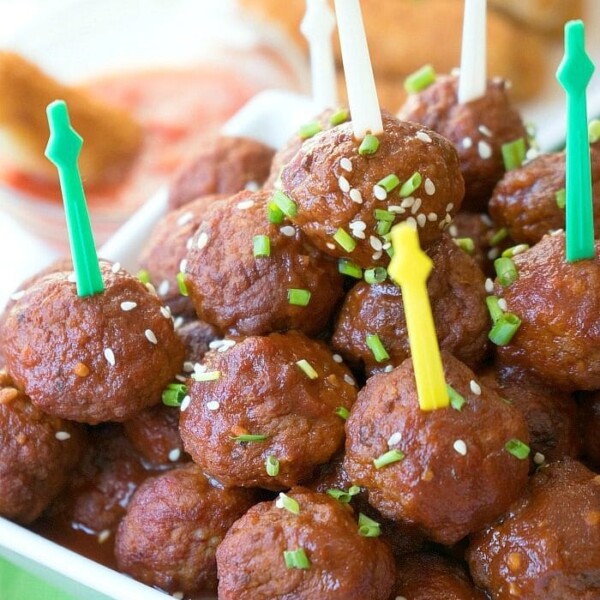 A hand picking up a glazed barbecue meatball with a toothpick from an instant pot dish, garnished with sesame seeds and chives.