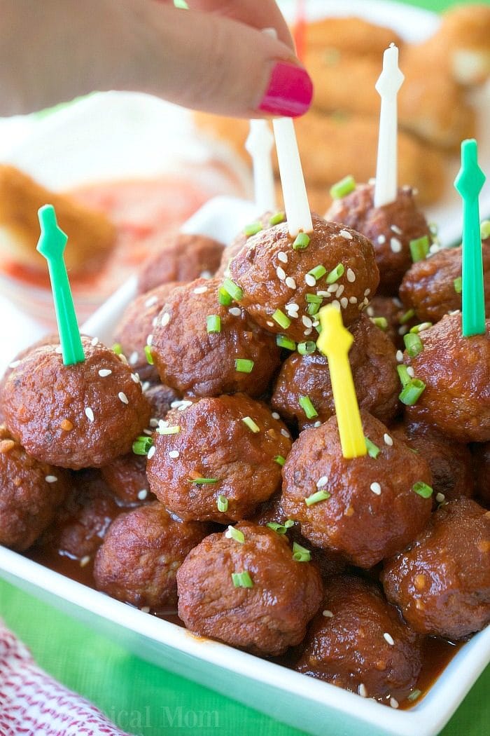 A hand picking up a glazed barbecue meatball with a toothpick from an instant pot dish, garnished with sesame seeds and chives.