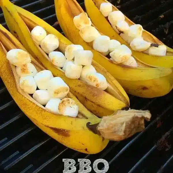 Three bananas are sliced open and filled with marshmallows and chocolate, then placed on a grill to create the ultimate banana s'mores. The text reads "BBQ Banana S'mores.