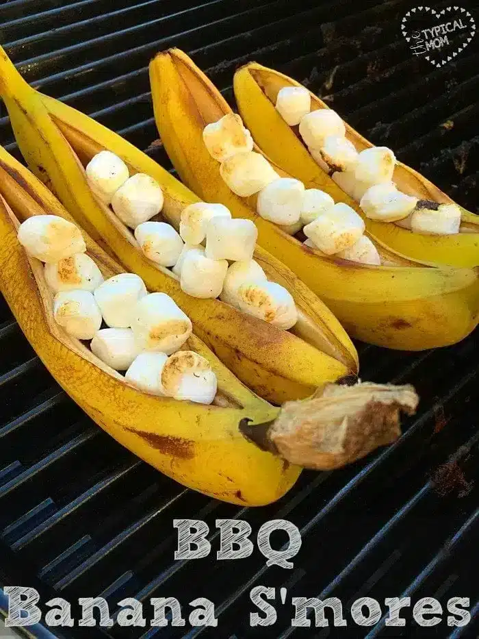 Three bananas are sliced open and filled with marshmallows and chocolate, then placed on a grill to create the ultimate banana s'mores. The text reads "BBQ Banana S'mores.