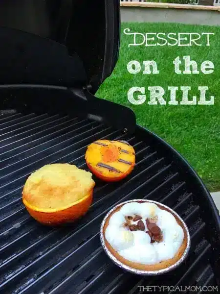 A grill with three decadent desserts: a grilled peach with char marks, a baked orange half, and a small pie with toasted marshmallows and chocolate pieces for banana s'mores. 