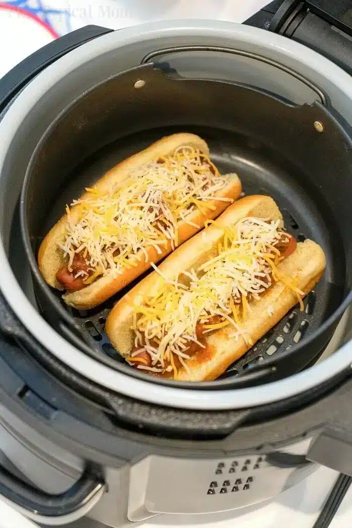 Two air fryer hot dogs with grated cheese on top sit in a basket, nestled in their buns side by side. The air fryer lid is visible at the edge of the image, capturing a perfect meal prep moment.