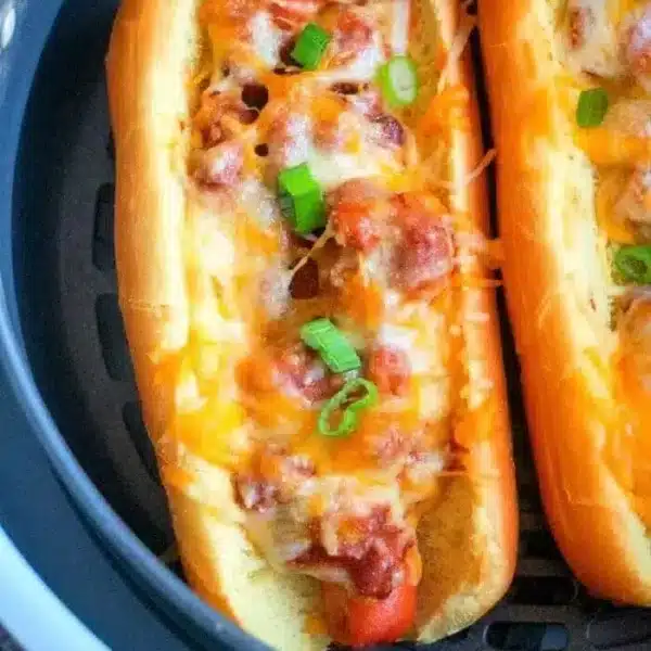 Two air fryer hot dogs topped with melted cheese, diced green onions, and chili sit enticingly in an air fryer basket. Nestled in soft buns, the filling overflows slightly. The background features a close-up of the air fryer and the savory hot dogs.