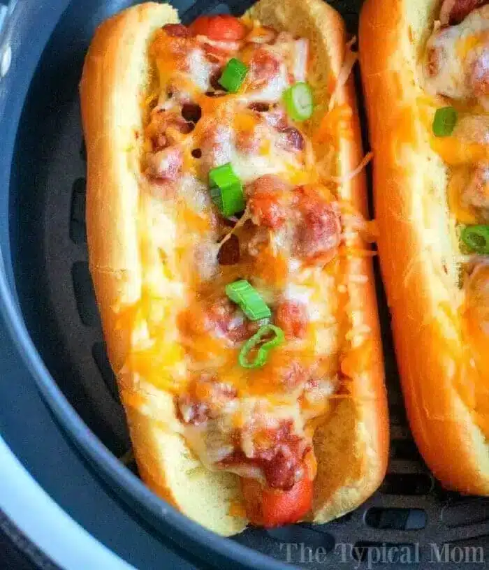 Two air fryer hot dogs topped with melted cheese, diced green onions, and chili sit enticingly in an air fryer basket. Nestled in soft buns, the filling overflows slightly. The background features a close-up of the air fryer and the savory hot dogs.