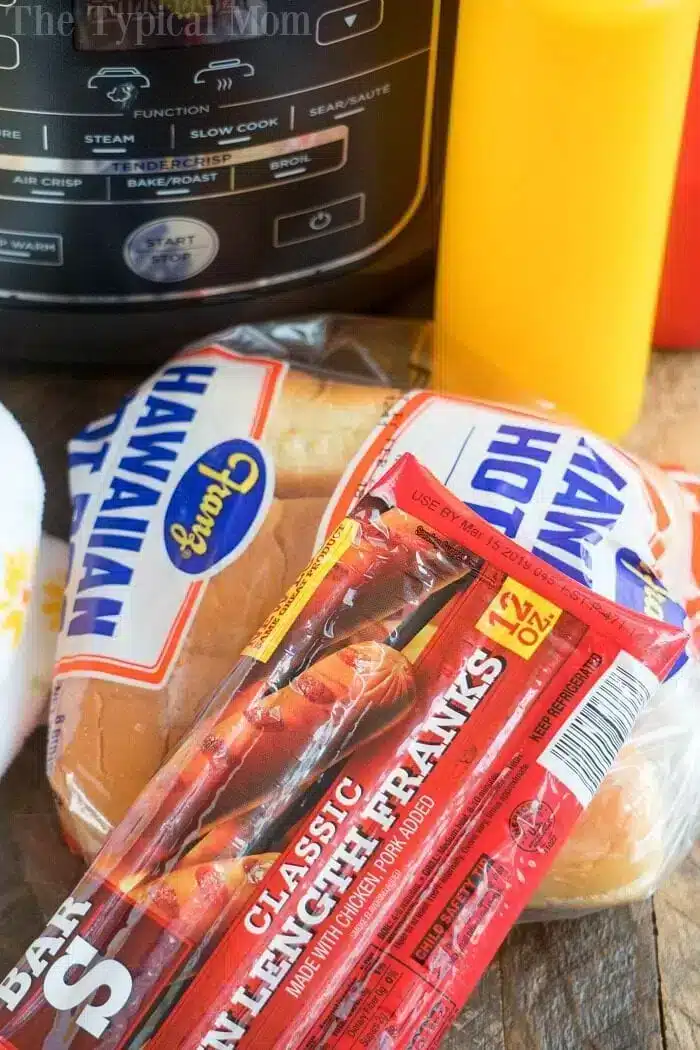 A package of classic lunch franks sits on a wooden surface next to a pack of Hawaiian hot dog buns. Behind them, an air fryer is ready for perfectly crispy hot dogs, and a yellow condiment bottle is partially visible.