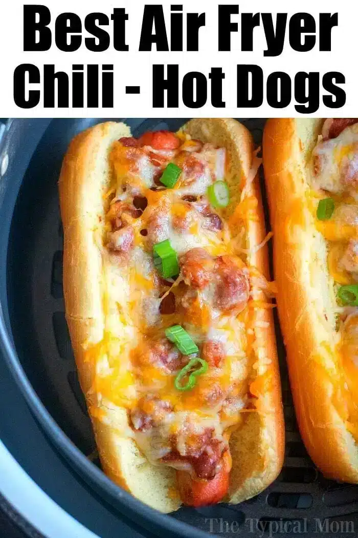 Two hot dogs in buns are topped with chili, melted cheese, and chopped green onions. The air fryer hot dogs are situated in an air fryer basket. The text at the top of the image reads, "Best Air Fryer Chili - Hot Dogs.