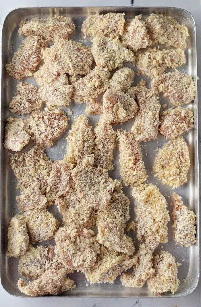 A baking tray filled with raw, breaded chicken pieces, reminiscent of air fryer popcorn chicken. The chicken is coated with breadcrumbs and arranged unevenly on the metal tray, ready for cooking.