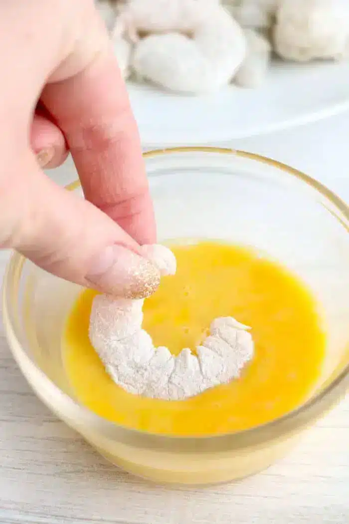 A hand is dipping a flour-coated shrimp into a bowl of beaten eggs. More flour-coated shrimp are visible in the background on a plate. The scene appears to be part of preparing bang bang shrimp, likely breading or battering them before cooking in an air fryer.