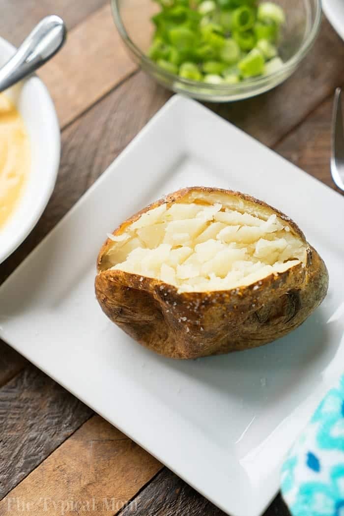 Easy air fryer baked potatoes. #airfryer #bakedpotatoes #ninjafoodi A fluffy air fryer baked potato on a white square plate, with a bowl of chopped green onions nearby on a wooden table.