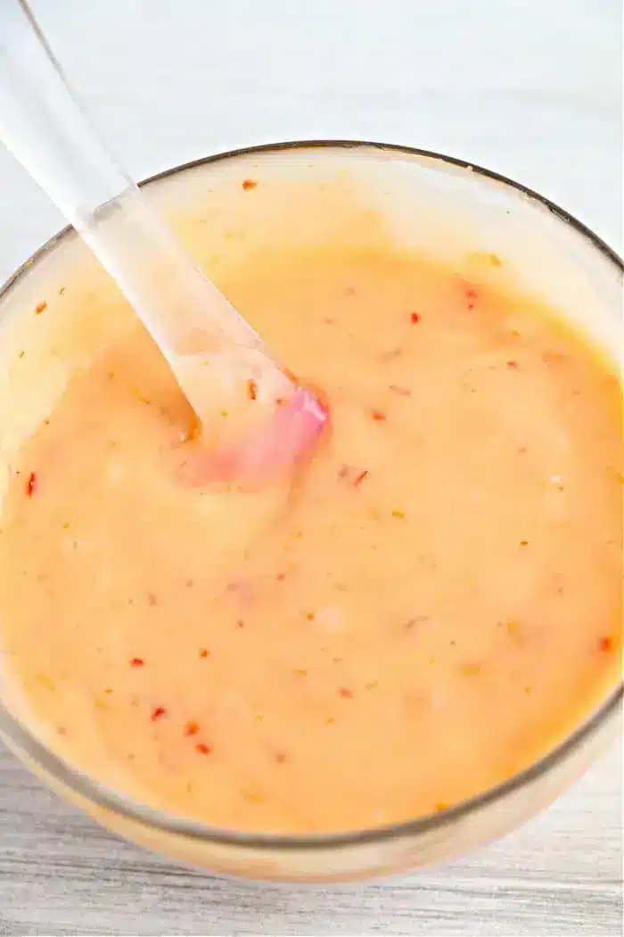 A clear bowl filled with a creamy orange sauce containing red flecks, reminiscent of the perfect dip for air fryer bang bang shrimp. A transparent spoon is partially submerged in the sauce, and the background is a white surface.