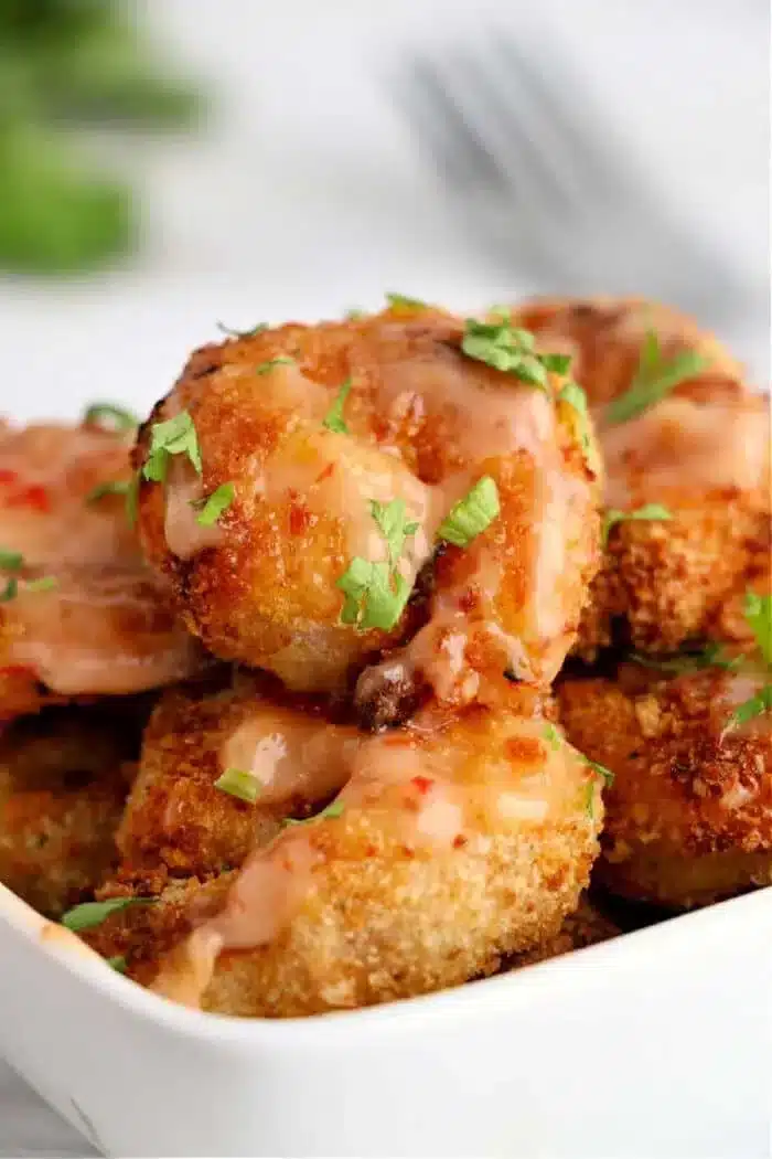 A close-up shot of a white dish filled with air fryer bang bang shrimp, topped with a drizzle of pink sauce and garnished with chopped green herbs. A softly blurred fork and greenery are visible in the background.