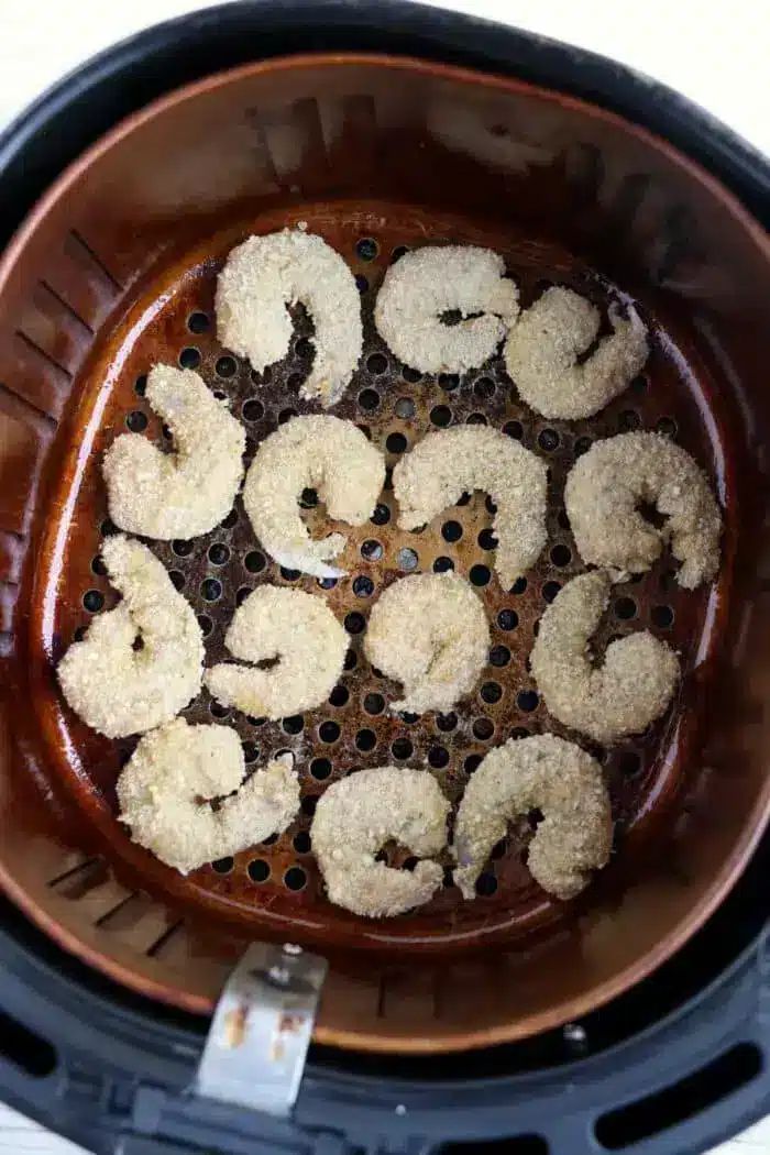 An air fryer basket containing twelve breaded bang bang shrimp arranged in a circular pattern. The shrimp are partially cooked, with a light golden-brown appearance. The basket has a perforated base.