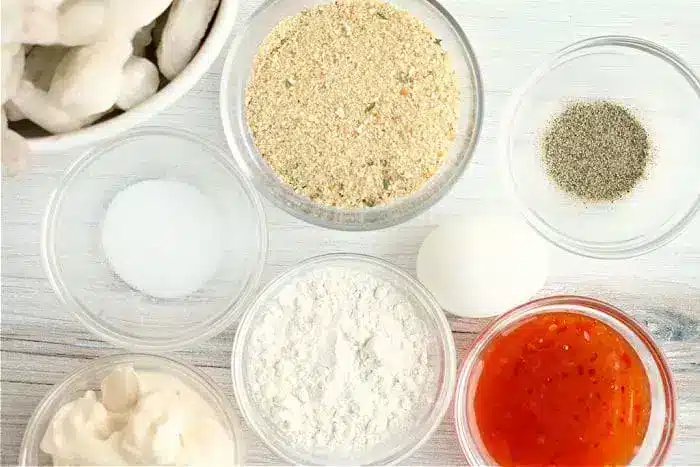 Ingredients arranged on a white surface: a small bowl of bread crumbs, black pepper, a white egg, flour, a red sauce for the bang bang shrimp, mayonnaise, salt, and a bowl of raw shrimp ready for the air fryer.