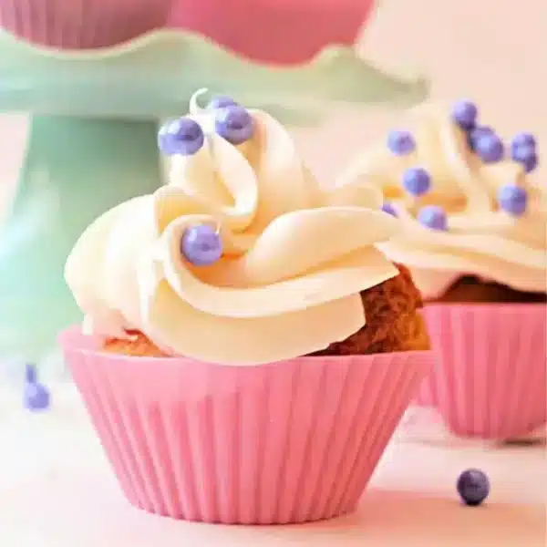 A cupcake with white frosting and purple candy pearls, made expertly in an air fryer, sits in a pink cupcake liner.