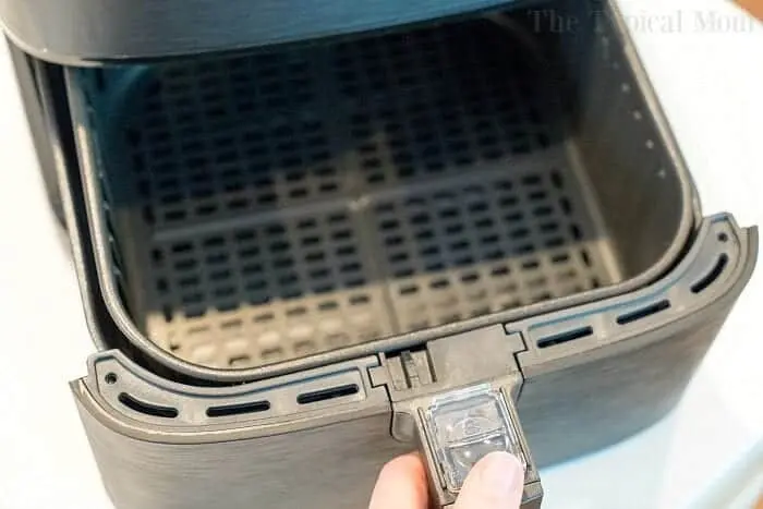 A close-up photo of a hand pulling out the basket of an open air fryer on a light-colored surface. The interior of the air fryer basket is empty and features a mesh design, perfect for cooking your favorite dishes like bang bang shrimp. The edge of the air fryer has a latch mechanism for securing the basket.