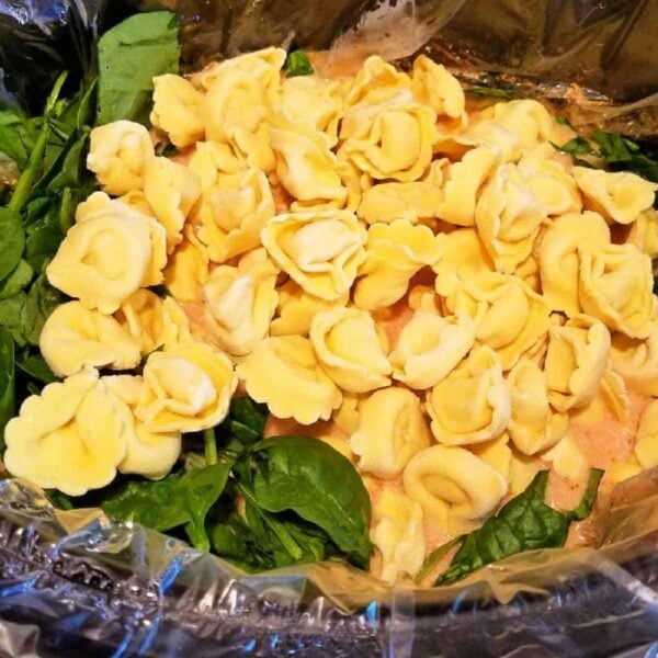 Uncooked tortellini pasta and fresh spinach in a slow cooker lined with a plastic liner, ready to be transformed into hearty crockpot tortellini soup.
