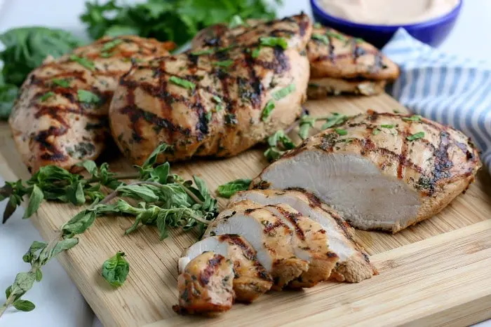 Grilled chicken breasts on a wooden cutting board, garnished with herbs and enhanced by the best fajita marinade. Several pieces are intact, while some are sliced. A small bowl of sauce is partially visible in the background, with fresh greens nearby.
