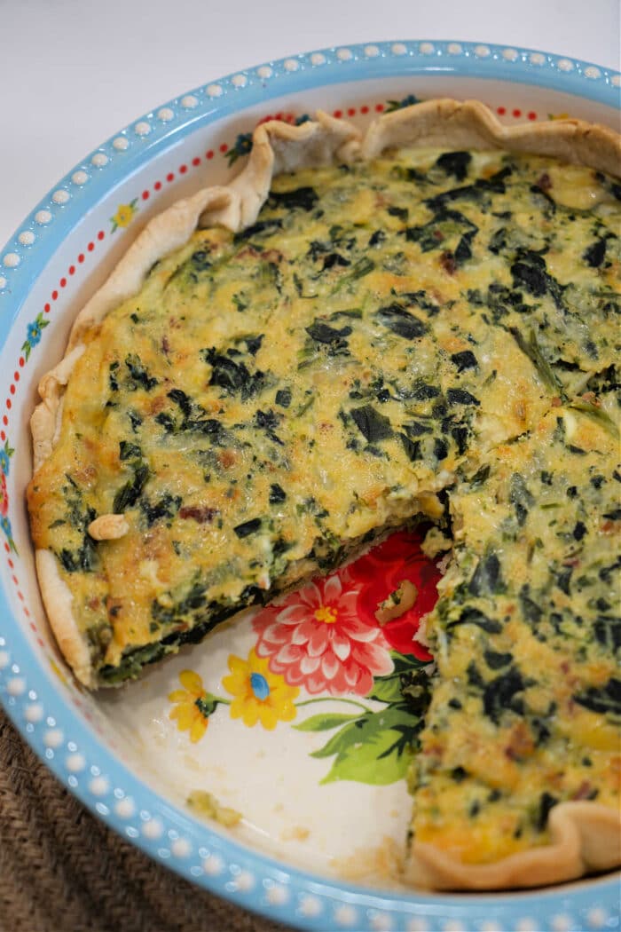 An easy spinach quiche recipe is showcased in a decorative pie dish with one slice missing, revealing the savory filling.