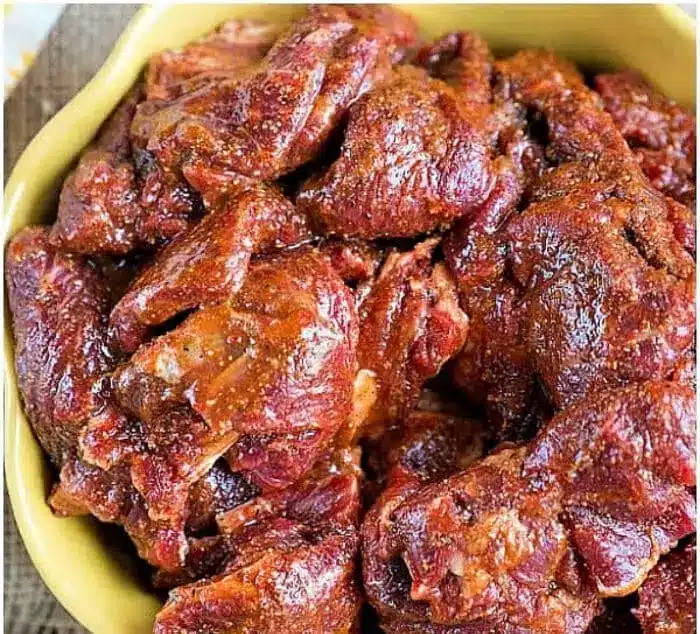 A bowl filled with raw, seasoned chunks of red meat, marinated with the best fajita marinade. The meat appears to be heavily coated with spices. The bowl is placed on a textured surface.