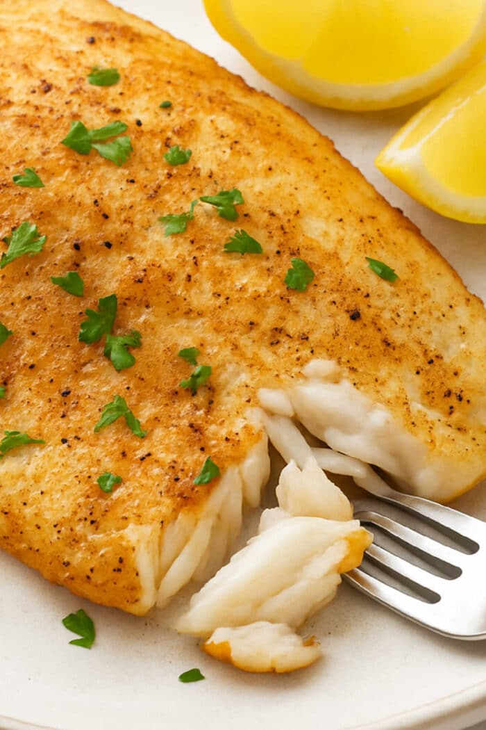 Golden, pan-seared fish fillet made from Frozen Tilapia Air Fryer, garnished with parsley and served with lemon wedges and a forkful of fish.