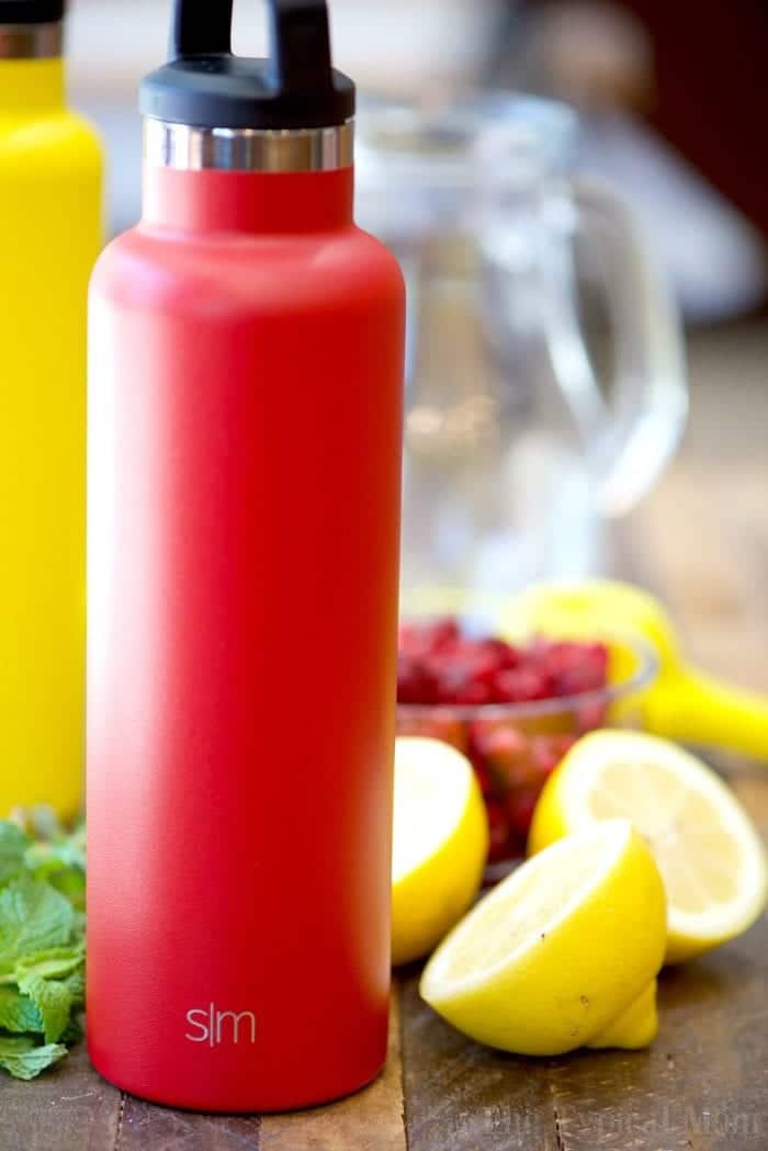 Red water bottle with lemons and a hint of homemade raspberry lemonade beside a glass pitcher in the background on a wooden table.