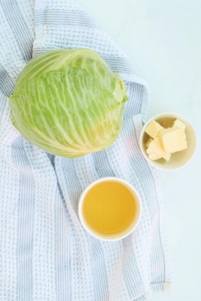 A head of cabbage, a bowl of butter, and a bowl of broth on a striped kitchen towel—perfect ingredients for preparing delicious Buttered Pressure Cooker Cabbage.