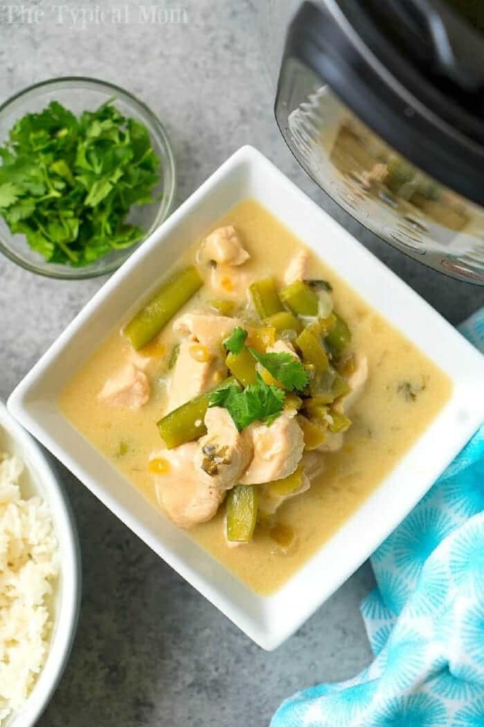 Pressure cooker coconut chicken with curry turned out amazing! A creamy Instant Pot chicken with vegetable dish served as a soup or on top of rice. Try our Thai style coconut chicken yourself and let us know what you think! #instantpot #pressurecooker #thai #indian #coconutchicken #instantpotrecipes #thetypicalmom #chicken #dairyfree #soup #curry Square bowl of creamy coconut chicken curry with green beans, next to rice, cilantro, and an Instant Pot.