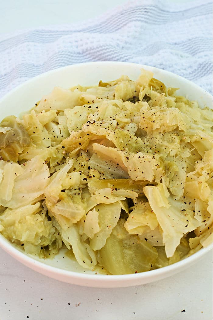 A bowl of Buttered Pressure Cooker Cabbage pieces, seasoned with black pepper, sits on a white surface next to a cloth.