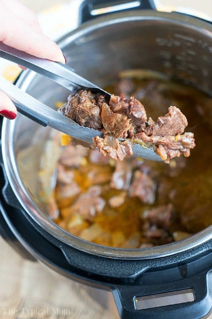 Tongs holding cooked beef chunks over an open Instant Pot with savory french onion pressure cooker roast stew inside.
