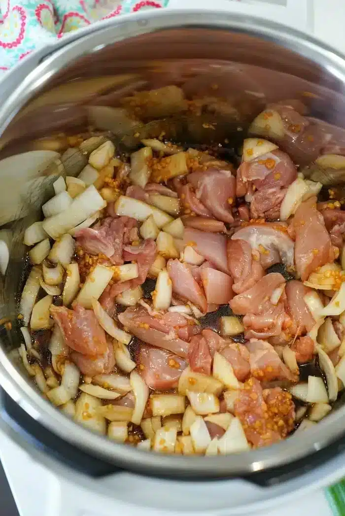 A metal pot on a stove contains uncooked diced chicken, chopped onions, and minced garlic. The ingredients are in the process of being sautéed, with some onions already starting to turn translucent, creating the base for a delicious Instant Pot Korean chicken dish.