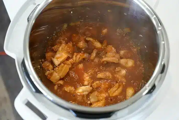 A close-up view of an Instant Pot containing chicken pieces simmering in a red tomato-based sauce. The dish, possibly Korean chicken, appears to be in the process of cooking, with visible steam and some chopped onions mixed in.