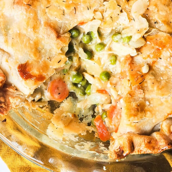 A freshly baked pot pie with a slice removed reveals a savory blend of leftover smoked chicken, peas, and carrots filling, set on a yellow cloth.