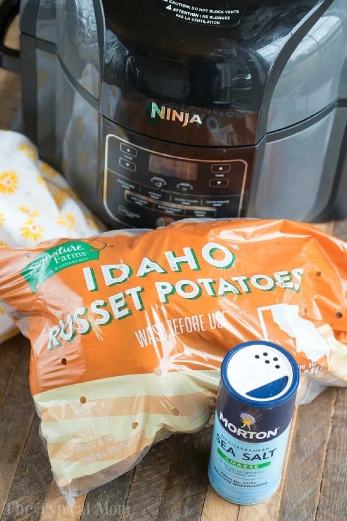 A Ninja air fryer, a bag of Idaho russet potatoes, and a container of sea salt on a wooden surface—everything you need for perfect air fryer baked potatoes.