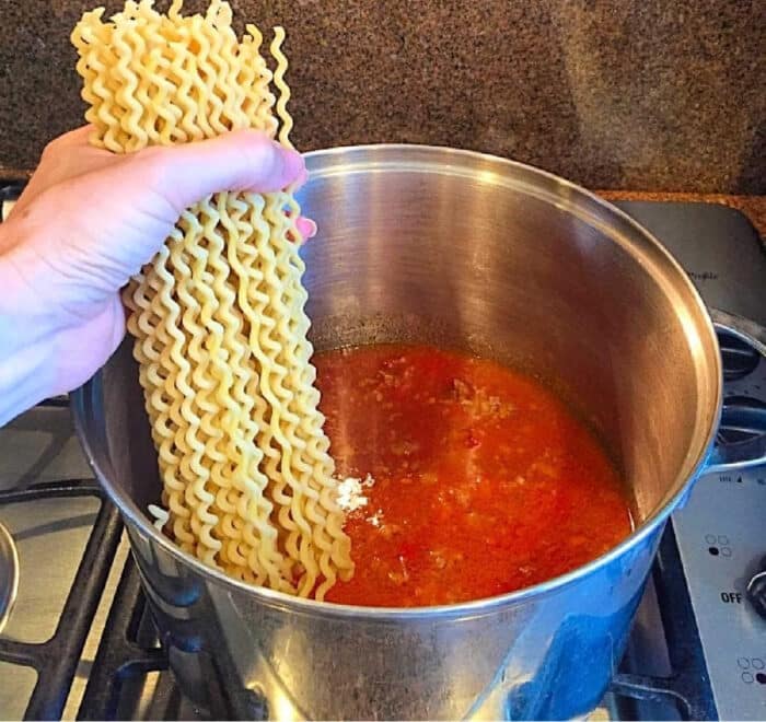 A hand holding dry curly pasta over a pot of red sauce on a stovetop, about to add the pasta for an easy one pot spaghetti meal.