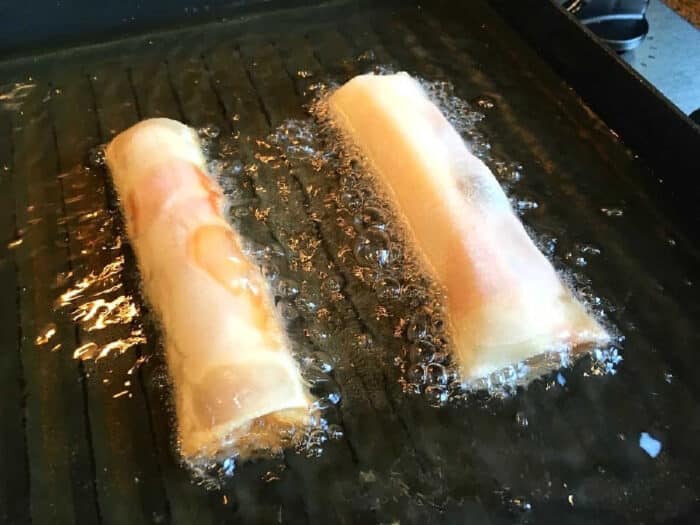 Two pizza egg rolls frying in hot oil with bubbles forming around them in a pan.