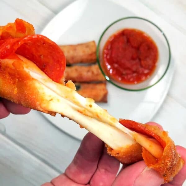 A hand pulls apart a cheesy pepperoni pizza stick with marinara sauce, while crispy pizza egg rolls and more sticks sit on a plate in the background.