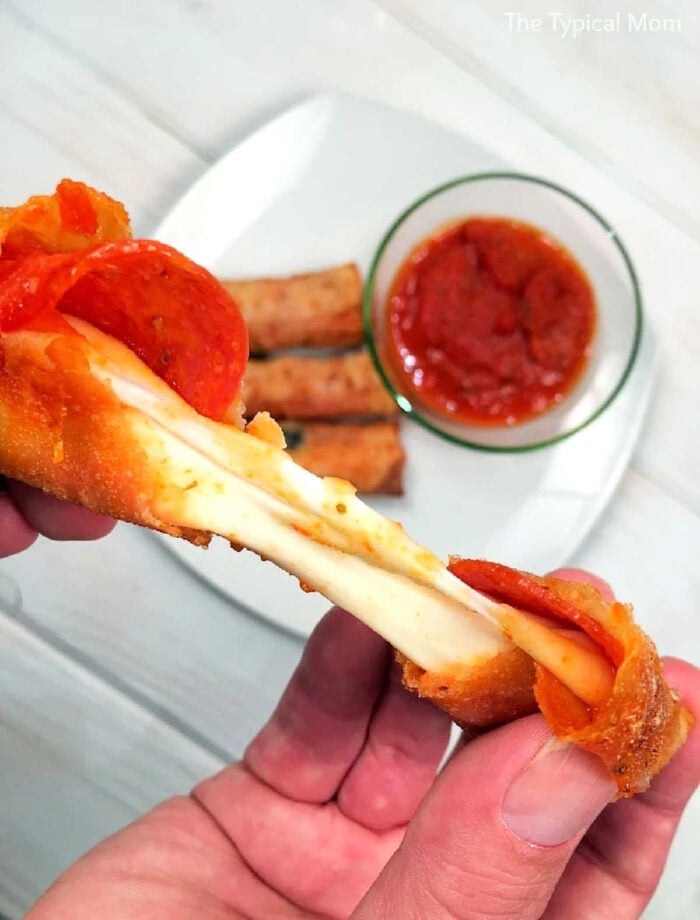 A hand pulls apart a cheesy pepperoni pizza stick with marinara sauce, while crispy pizza egg rolls and more sticks sit on a plate in the background.