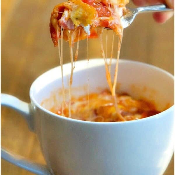 A fork lifts cheesy pizza in a mug mug cake from a white mug on a wooden table.