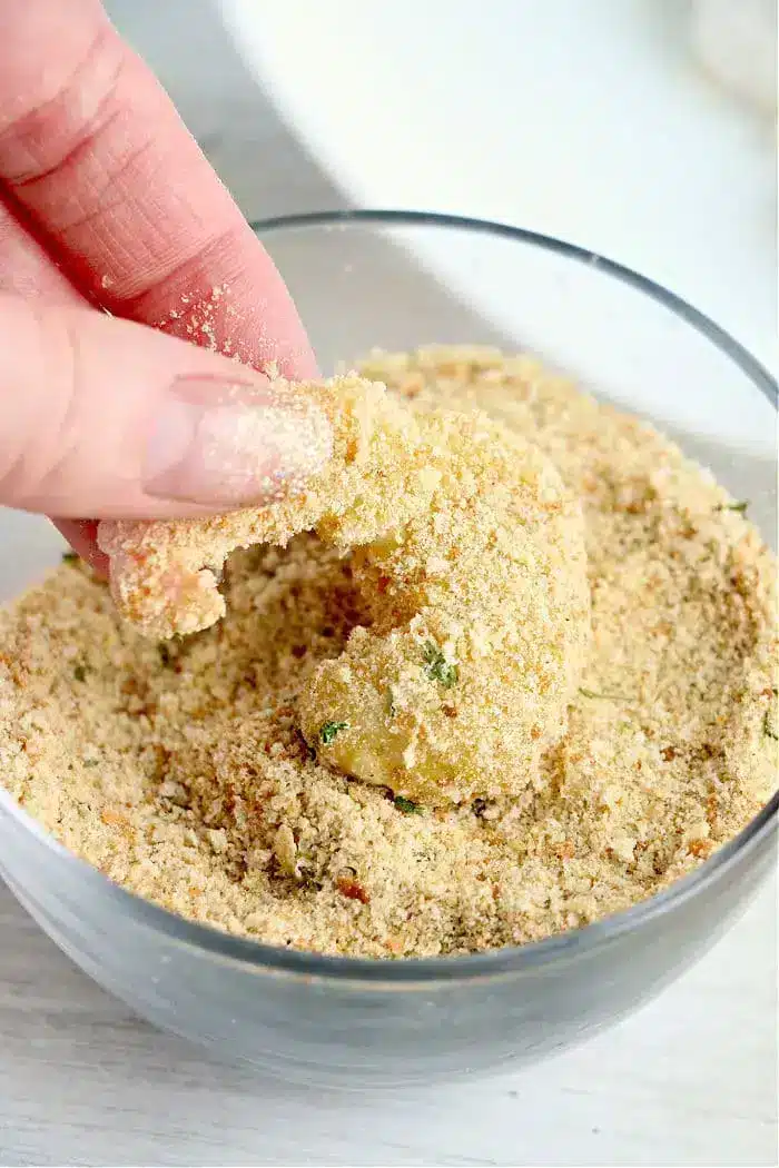 A hand is shown dipping a piece of food, possibly bang bang shrimp, into a bowl of seasoned bread crumbs. The bread crumbs appear coarse and contain various herbs. The partly coated shrimp will soon be ready for the air fryer, promising a crispy and flavorful delight.