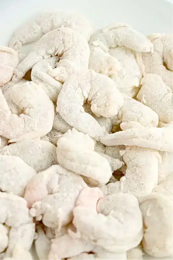 A close-up view of a pile of raw shrimp covered in a layer of flour or breading on a white background, ready to be transformed into crispy air fryer bang bang shrimp.