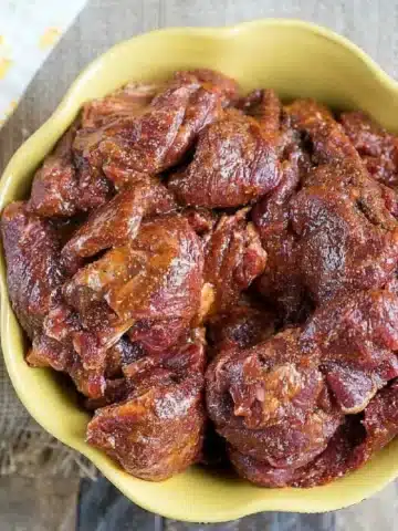 A bowl filled with seasoned pieces of raw meat, placed on a wooden surface. The reddish-brown color suggests the use of the best fajita marinade, infused with spices. The light brown bowl has a decorative edge, enhancing its rustic appeal.