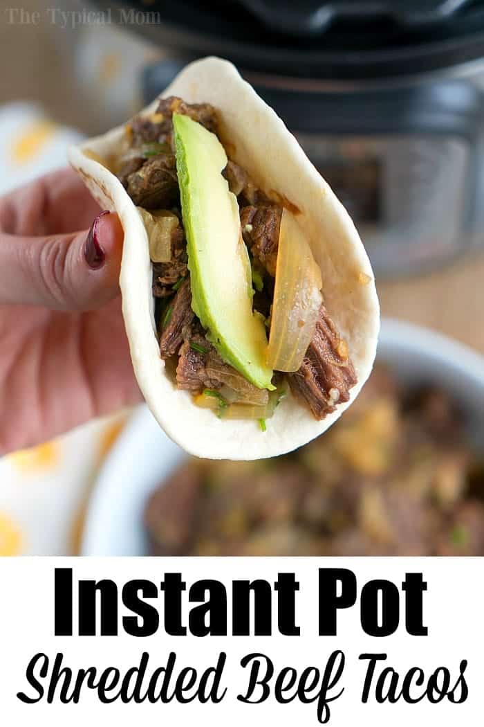 French onion pressure cooker roast is tender with tons of flavor. Perfect Instant Pot shredded beef tacos for dinner even your kids will rave about. #instantpotrecipes #instantpot #roast #frenchonion #tacos #shreddedbeef #ninjafoodirecipes #crockpotexpressrecipes #pressurecookerrecipes Hand holding a soft taco filled with french onion pressure cooker roast, shredded beef, onions, and avocado, with Instant Pot in background.