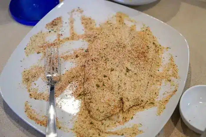 A piece of air fryer tilapia is covered in a seasoned breadcrumb mixture on a white plate. A silver fork rests on the side of the plate, and there is a small empty white bowl and a blue lid in the background.