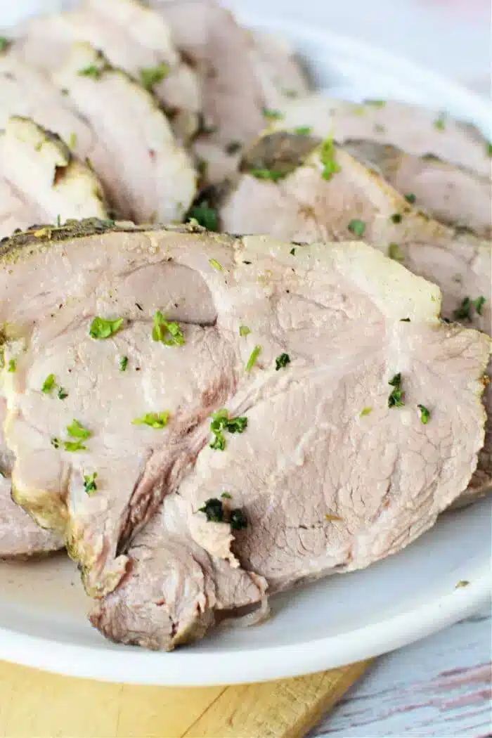 Slices of roasted pork, straight from the slow cooker, are garnished with chopped herbs and arranged on a white plate. The meat appears tender and well-cooked.