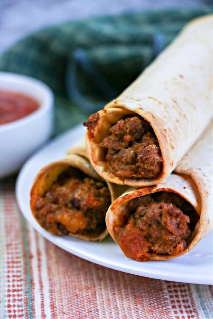 Three beef-filled air fryer flautas stacked on a plate with a bowl of salsa in the background.