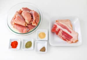 Bacon wrapped chicken thighs and raw bacon with spices rest enticingly on a pristine white surface.