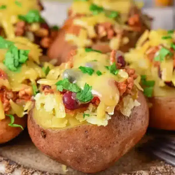 Three baked red potatoes are topped with melted cheese, chili with beans, and garnished with sprigs of fresh parsley, served on a wooden plate with a blurred background featuring condiments.