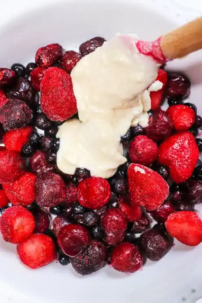 A bowl of assorted berries, including strawberries, blueberries, and blackberries, topped with a dollop of creamy batter reminiscent of a Bisquick cobbler. A wooden spoon is partially dipped into the mixture.